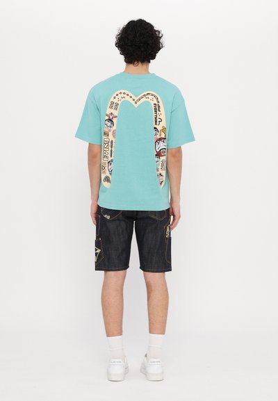 Τυρκουάζ βαμβακερό μπλουζάκι με μεγάλη γραφιστική σχεδίαση στην πλάτη, συνδυασμένο με σκούρες τζιν βερμούδες με ραφές και λεπτομέρειες.
