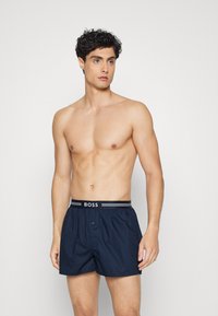 Boxer shorts bleu marine avec une ceinture élastique arborant l'inscription "BOSS" en blanc, dotés d'une fermeture à bouton et d'une coupe ample.