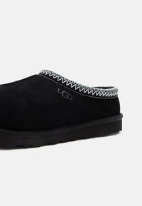 Chaussure noire en daim à enfiler avec un design texturé, couture blanche le long du col et semelle en caoutchouc noir. Logo UGG visible.