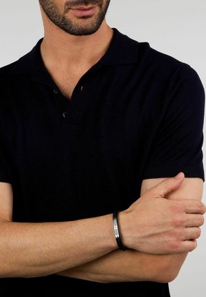 Mann trägt ein kurzärmeliges schwarzes Polohemd und ein schwarzes Lederarmband mit silberner Schnalle, die Arme auf Brusthöhe verschränkt.