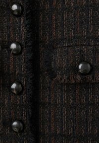 Teksturowana ciemna tkanina z wzorem tkaniny z pionowymi paskami w odcieniach czerni i brązu. Widać pięć błyszczących czarnych guzików.