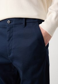 Navy blue cotton trousers with a smooth texture, featuring a button closure and side pockets. The waistband is partially covered by a cream ribbed sweater.