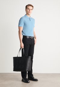 Cămașă polo de un albastru deschis, blugi negri și pantofi negri. Ține o geantă mare din piele neagră cu text embossat „HUGO”. Texturi fine.