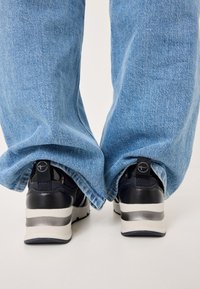 Sneakers con tomaia in pelle nera e dettagli argentati, abbinati a jeans in denim azzurro chiaro e larghi, visibili da dietro.