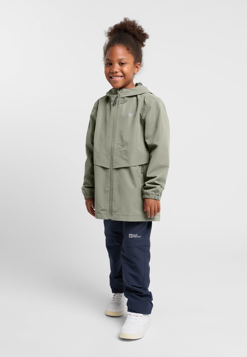 Giacca impermeabile verde chiaro con cappuccio, chiusura a zip, polsini elasticizzati. Indossata con pantaloni blu navy e sneakers bianche, in piedi su uno sfondo neutro.