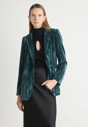 Teal velvet blazer with a textured finish, single button closure, and two front pockets, worn over a black high-neck top and skirt.