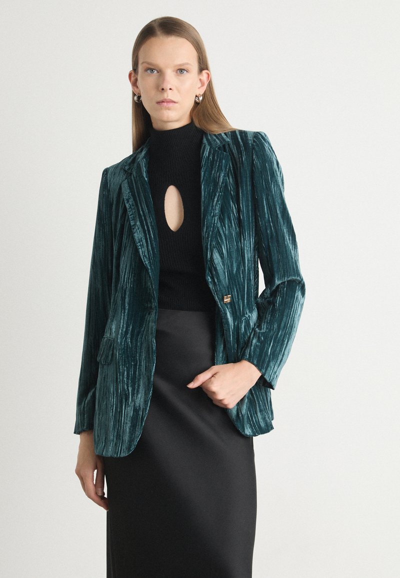 Teal velvet blazer with a textured finish, single button closure, and two front pockets, worn over a black high-neck top and skirt.