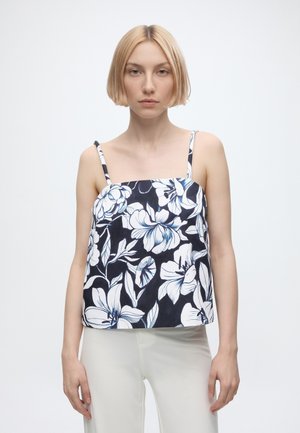 Femme aux cheveux blonds coiffés en carré, portant un top à fines bretelles bleu marine et blanc à motifs floraux ainsi qu’un pantalon clair, debout devant un fond uni.