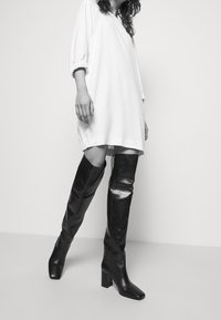 Svarta läderstövlar över knät med fyrkantig klack. Slät textur, glansig yta, tillsammans med en vit, löst sittande klänning med puffärmar.