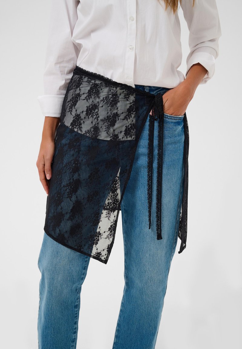 Femme portant un jean bleu et une chemise blanche avec un foulard en dentelle noire noué autour de la taille.