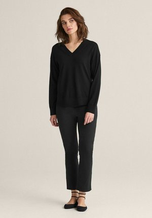 Sort v-hals sweater med lange ærmer, nænsomt parret med tætsiddende sorte bukser og sorte flade sko. Glat tekstur, afslappet design, minimal hardware.