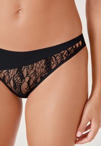 Perizoma in pizzo nero con un design in pizzo trasparente sui lati, completato da una vita nera opaca e una texture morbida.
