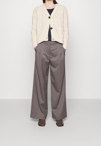 Cardigan de punto trenzado crema con botones negros sobre una blusa azul marino, combinado con pantalones de pierna ancha gris y botas de cuero marrón.