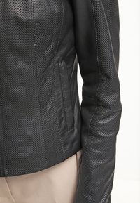 Svart läderjacka med perforerad textur, skräddarsydd design och sidofickor. Slät yta med åtsittande ärmar.