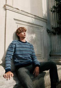 Strickpullover in Blautönen mit strukturierten Mustern, Rundhalsausschnitt und marineblauen Bündchen, kombiniert mit grauen Wollhosen, sitzend an einer Steinmauer.