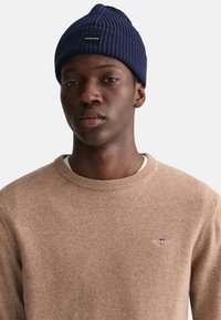 Beige gestrickter Pullover mit Rundhalsausschnitt und kleinem Logo-Detail. Marineblauer Rippstrick-Mütze mit einem Logo-Patch auf der Vorderseite.