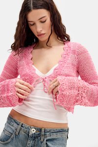Cardigan de punto rosa con bordes en forma de concha, de frente abierto y mangas acampanadas, superpuesto a una camiseta de tirantes blanca y combinado con jeans de denim.
