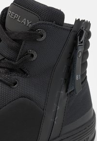 Černá kotníková teniska z texturované látky a gumy. Má nášivku s logem, šněrování vpředu a boční zip s visačkou označenou '81'.