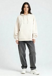 Vrouw met lang donker haar, gekleed in een oversized beige hoodie, losse donkergrijze spijkerbroek en witte sneakers, staande tegen een lichte achtergrond.