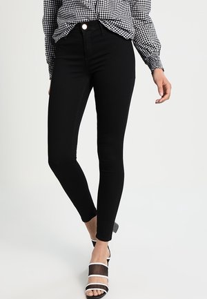 Femme portant un jean skinny noir taille haute, une chemise à carreaux noire et blanche, et des talons ouverts noirs avec des brides blanches.