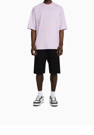 Mann trägt ein locker sitzendes helllila T-Shirt, schwarze Knie kurze Shorts, weiße Socken und schwarz-weiße Turnschuhe, steht vor weißem Hintergrund.