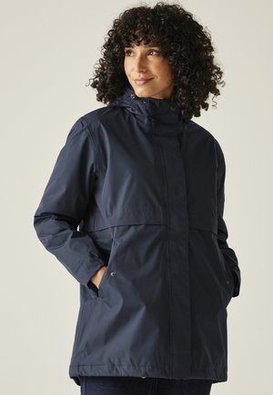 Kobieta z kręconymi włosami, ubrana w ciemnoniebieską kurtkę przeciwdeszczową z kapturem, z rękami w kieszeniach, patrząca na bok na jednolitym tle.