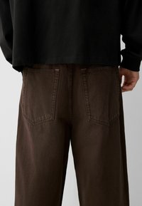 Smeđi traperice sa strukturiranim krojem, s dvije stražnje džepove s vidljivim šavovima. Materijal izgleda čvrsto s suptilnom teksturom.