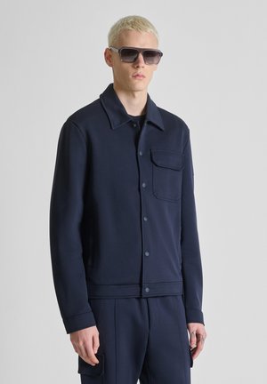 Jonge man met kort blond haar, draagt een oversized donkere zonnebril, een marineblauwe jas met knopen en een borstzak, en bijpassende marineblauwe broek.