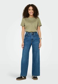 Oliven grøn t-shirt med øjenudskæring, parret med højtaljede, vide blå denimjeans og sorte sneakers med en beige sål.