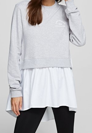 Kobieta ubrana w jasnoszarą bluzę z białym nadrukiem logo na białej długiej koszuli, w połączeniu z czarnymi legginsami, stoi na neutralnym tle.