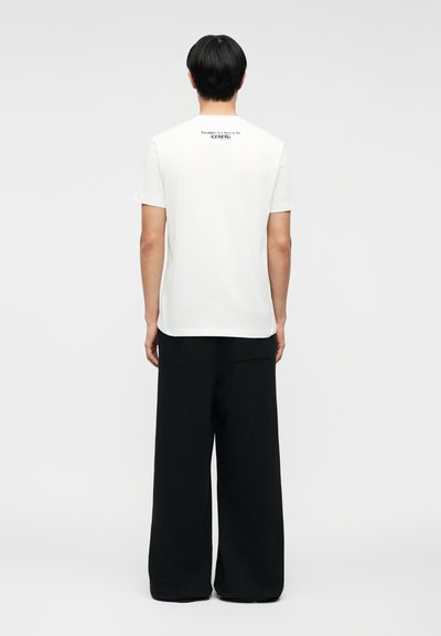 Άτομο με κοντά μαύρα μαλλιά στέκεται με την πλάτη γυρισμένη φορώντας λευκό κοντομάνικο πουκάμισο με μικρό κείμενο στο πάνω μέρος της πλάτης και μαύρο παντελόνι φαρδύ.