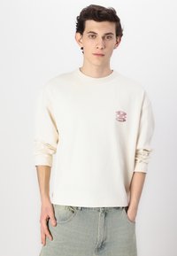 Camisa de manga larga crema con cuello redondo, que presenta un parche de logo burdeos en el lado izquierdo del pecho y un corte relajado, combinada con jeans azul claro.