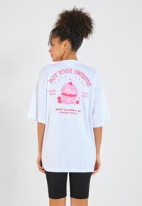 White oversized t-shirt featuring a pink graphic of a cupcake with text, and short sleeves. Worn with black bike shorts.