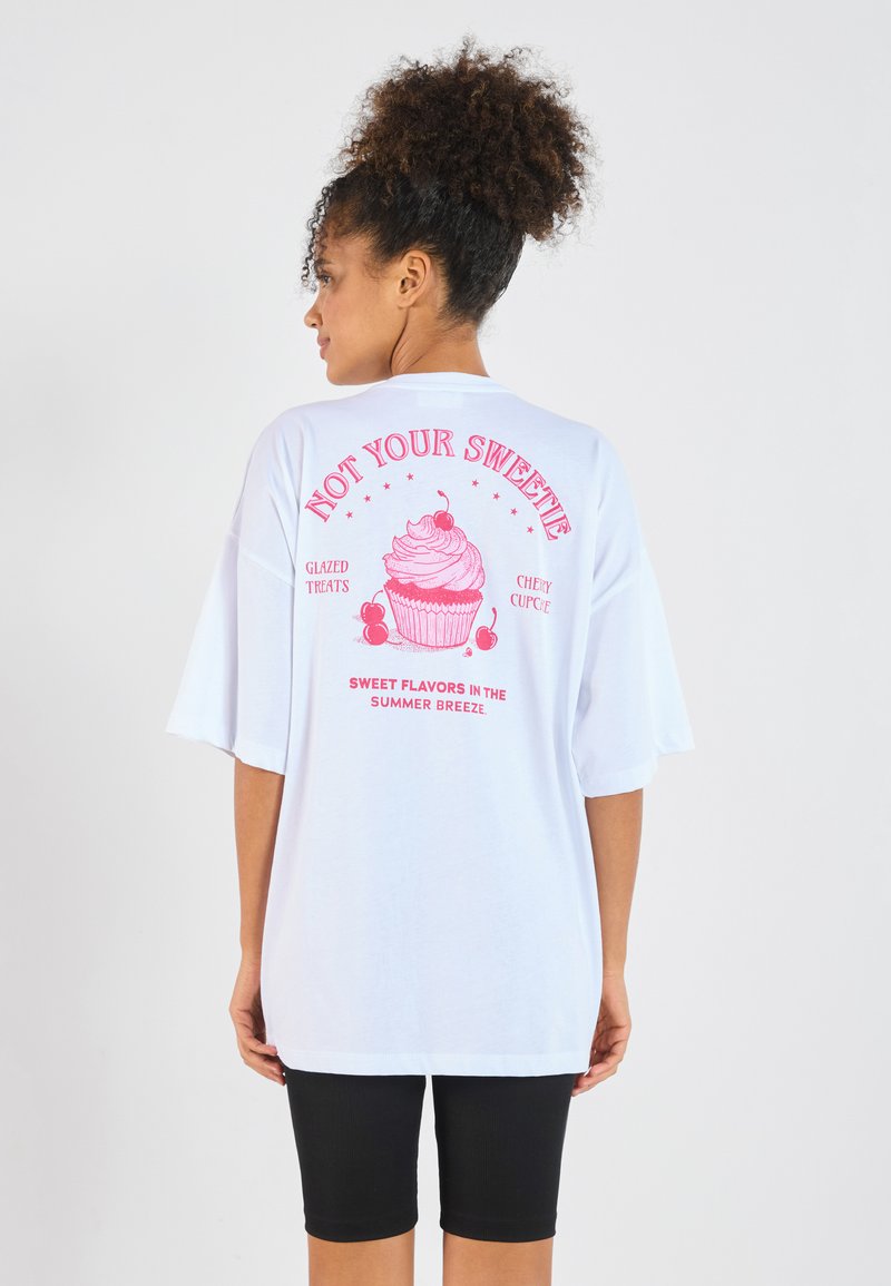Vit oversized t-shirt med en rosa grafisk bild av en cupcake med text, samt korta ärmar. Bärs tillsammans med svarta cykelshorts.