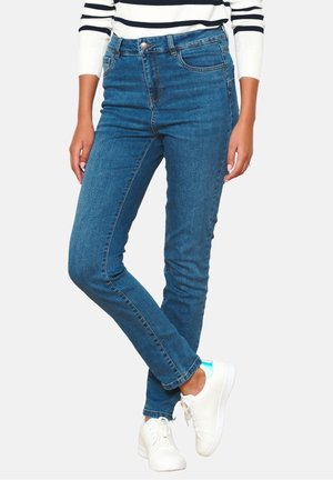 Person, die blaue Skinny-Jeans, weiße Sneakers mit blauen Details und ein weißes Langarmshirt mit schwarzen horizontalen Streifen trägt.