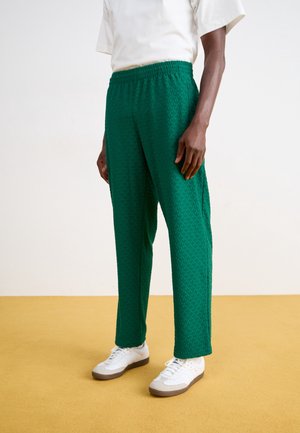 Mężczyzna ubrany w zielone spodnie o fakturze, białe trampki i białą koszulę z krótkim rękawem stoi na żółtej podłodze na tle jednolitej białej ściany.