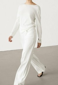 Haut en tricot à manches longues blanches associé à un pantalon large blanc, présentant une texture côtelée et une coupe décontractée. Bracelet à maillons en argent au poignet.