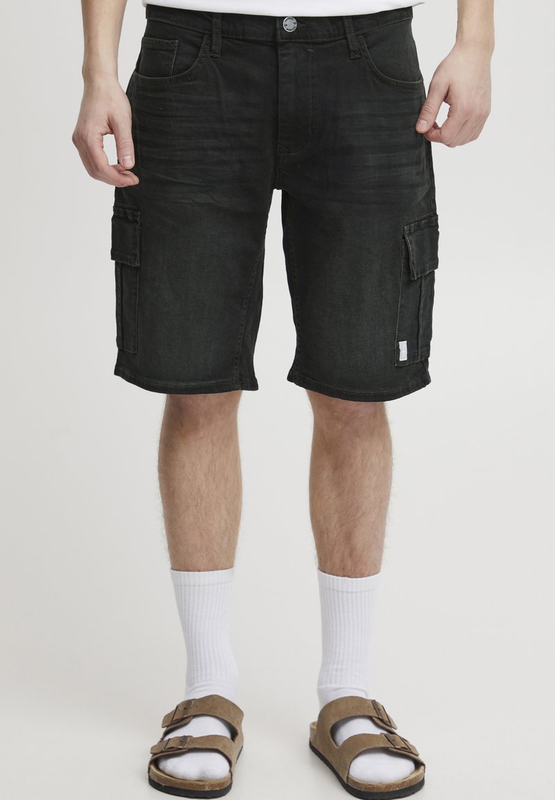Pantaloni cargo neri in denim con multiple tasche, vestibilità ampia e una texture sbiadita. Abbinati a calzini bianchi e sandali marroni.