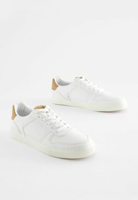 Witte sneakers van glad synthetisch materiaal, met een beige hakaccent, platte veters en een low-top ontwerp met zichtbare stiksels.