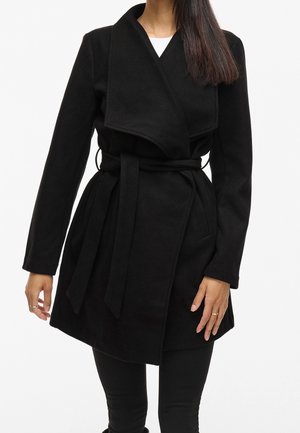 Vrouw draagt een zwarte wikkeljas met ceintuur en brede kraag over een zwarte broek, tegen een effen witte achtergrond.