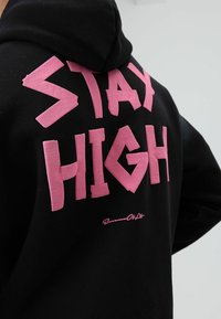 Zwarte hoodie met grote, roze grafische tekst "STAY HIGH" op de achterkant. De tekst is vetgedrukt en heeft een gestructureerde uitstraling.