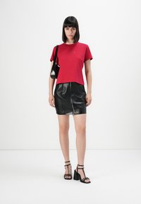 Donna con una maglietta rossa a maniche corte, una mini gonna in pelle nera con cerniera frontale, una borsa a spalla nera e sandali neri con tacco alto in piedi.