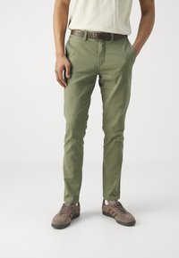Chinos verde claro de algodón, con un corte slim, bolsillos delanteros y un cinturón de cuero marrón. Los zapatos son marrones con acentos grises.
