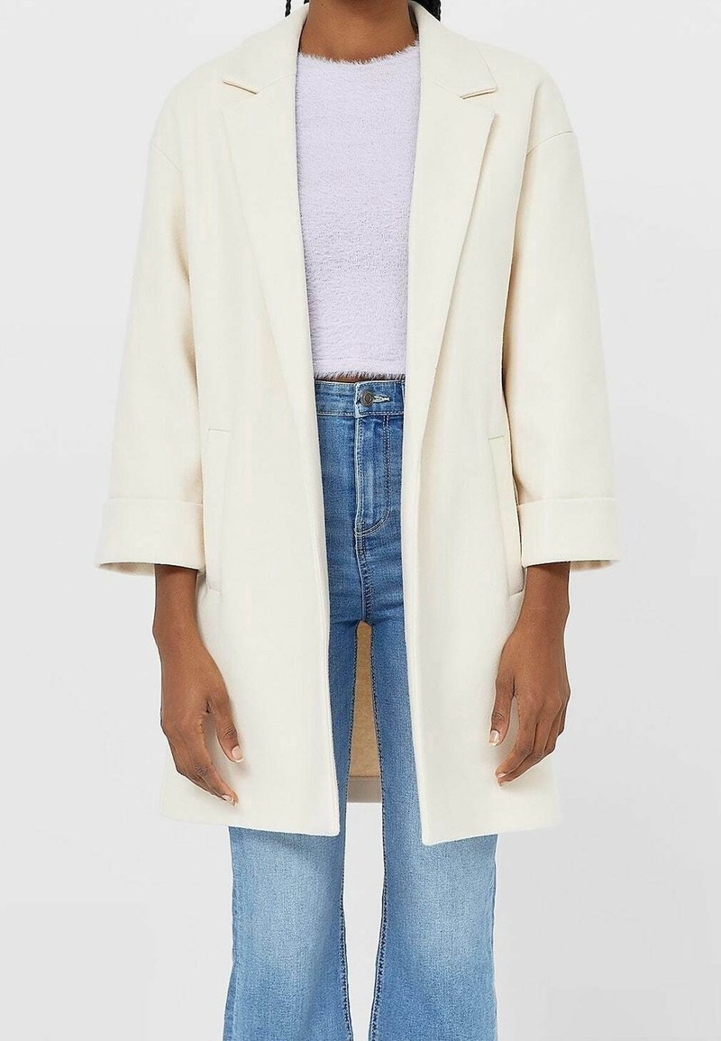 Cream oversized blazer with notched lapels, two front pockets, and rolled cuffs, paired with a light textured top and blue flared jeans.