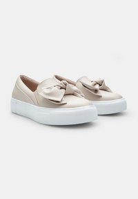 Löysi beige kengät, joissa on suuri satiininen solmu edessä, varustettuna tekstuurilla valkoisella kumipohjalla ja sileällä ylämateriaalilla.