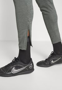 Pantalones deportivos grises con detalles de cremallera lateral, combinados con zapatillas Nike negras que tienen acentos blancos y un diseño de malla. Calcetines negros visibles.