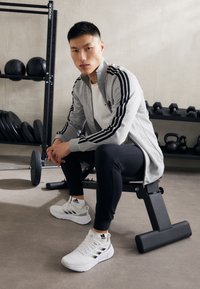 Siva atletska jakna z črnimi črtami, bela majica, črne legice in bele teniske. Fitnes okolje, uteži v ozadju.
