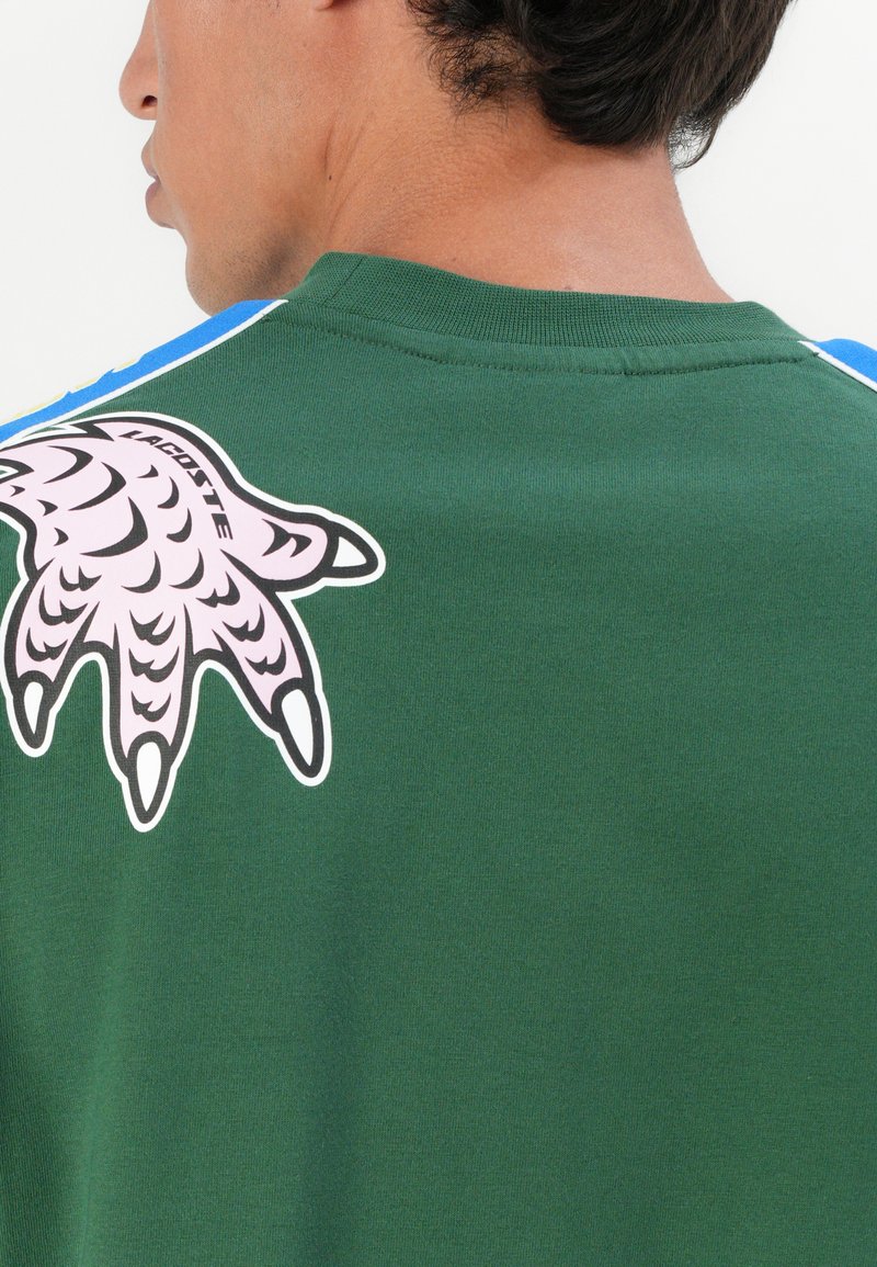 Zielona bluza z dużym różowym wzorem pazura na plecach, teksturowana tkanina i niebieskie akcenty na ramionach.