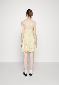 Vestido floral amarelo com alças finas, design em camadas e comprimento até a metade da coxa. Usado com sapatos pretos e meias brancas, visto pela parte de trás.