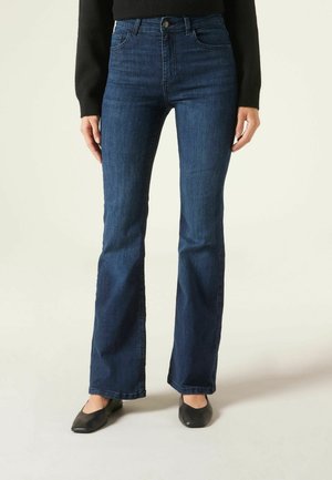 Femme portant un jean bootcut bleu foncé, un haut à manches longues noir et des chaussures plates noires, debout devant un fond clair uni.